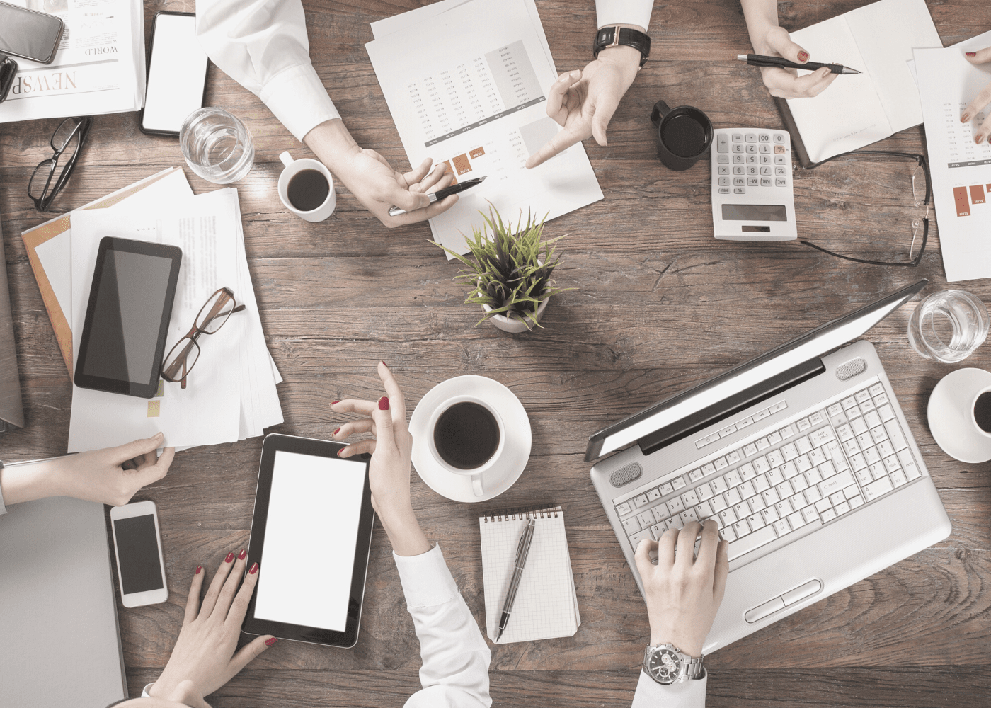 Bird-eye view of a table with people's hands working with coffee, papers, pens and other office supplies scattered